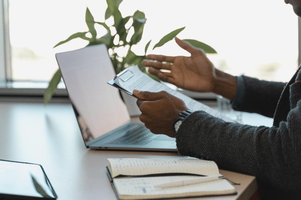 Professional reviewing documents and gesturing toward a clipboard beside a laptop on a desk